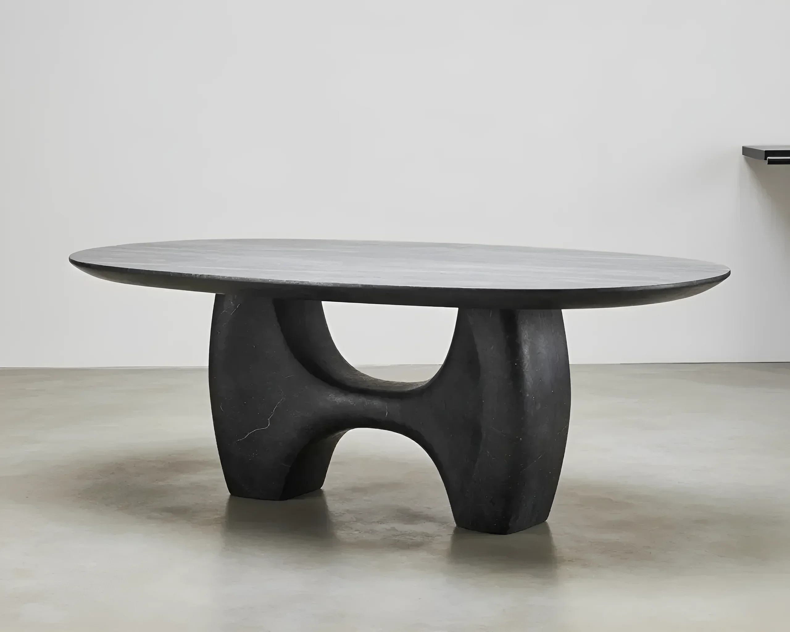 Black oval coffee table with a unique sculptural base resembling two curved arches, standing on a concrete floor.