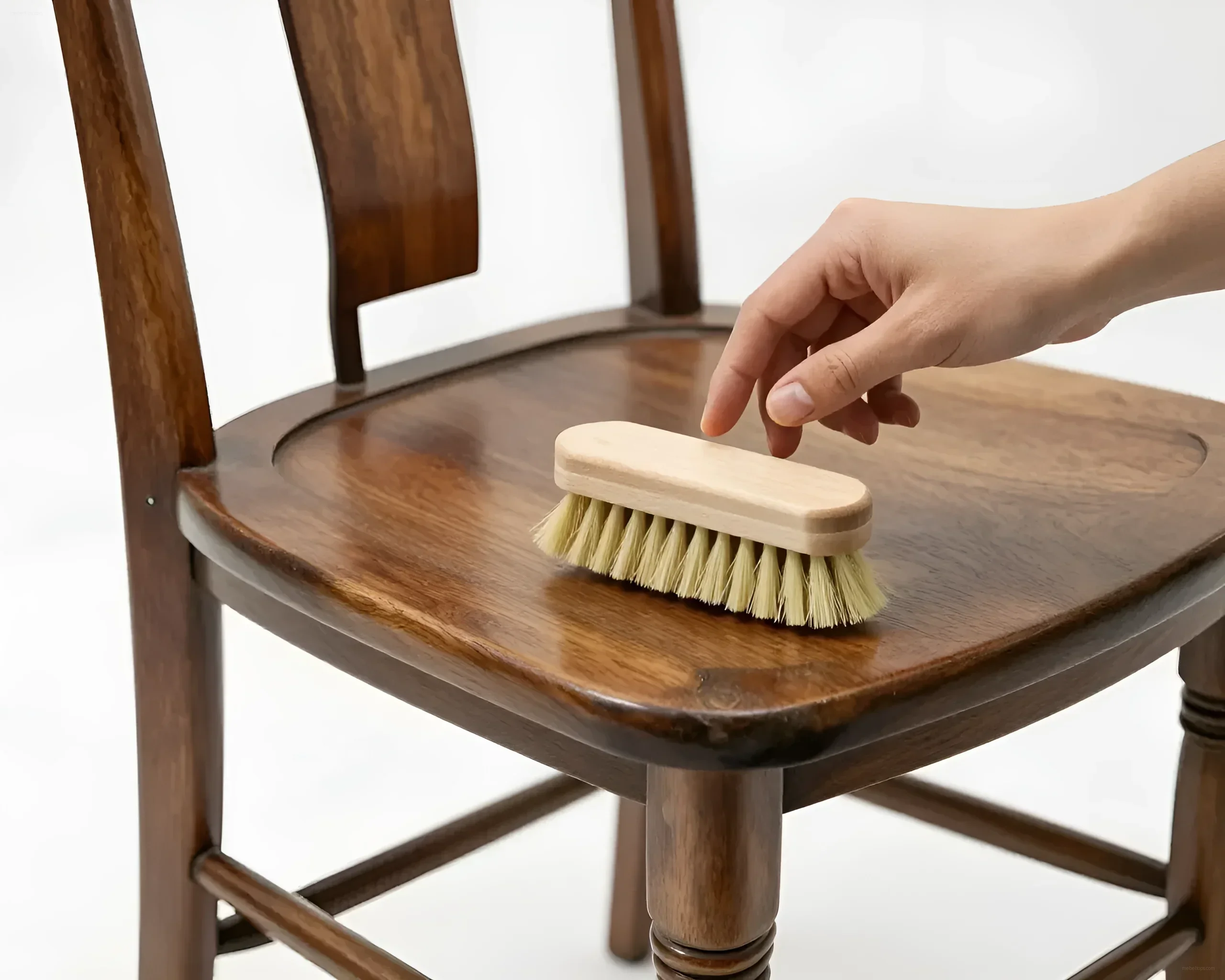 A person's hand with a soft furniture brush gently cleans the seat of a wooden antique chair, highlighting its natural texture and shine.