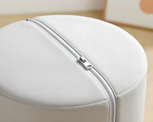 Крупный план белого круглого пуфа для детской комнаты с блестящей металлической молнией, проходящей по боковой поверхности.