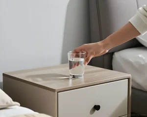 Рука ставит стакан воды на светлую прикроватную тумбу с ящиком