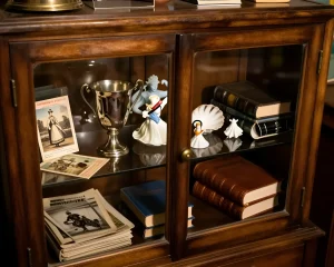 Красиво оформленная деревянная витрина со стеклянными дверцами, внутри — серебряный кубок, фарфоровые фигурки, ракушки, старые книги и открытки. Идеальный пример эстетичного хранения летних воспоминаний и сувениров из путешествий.